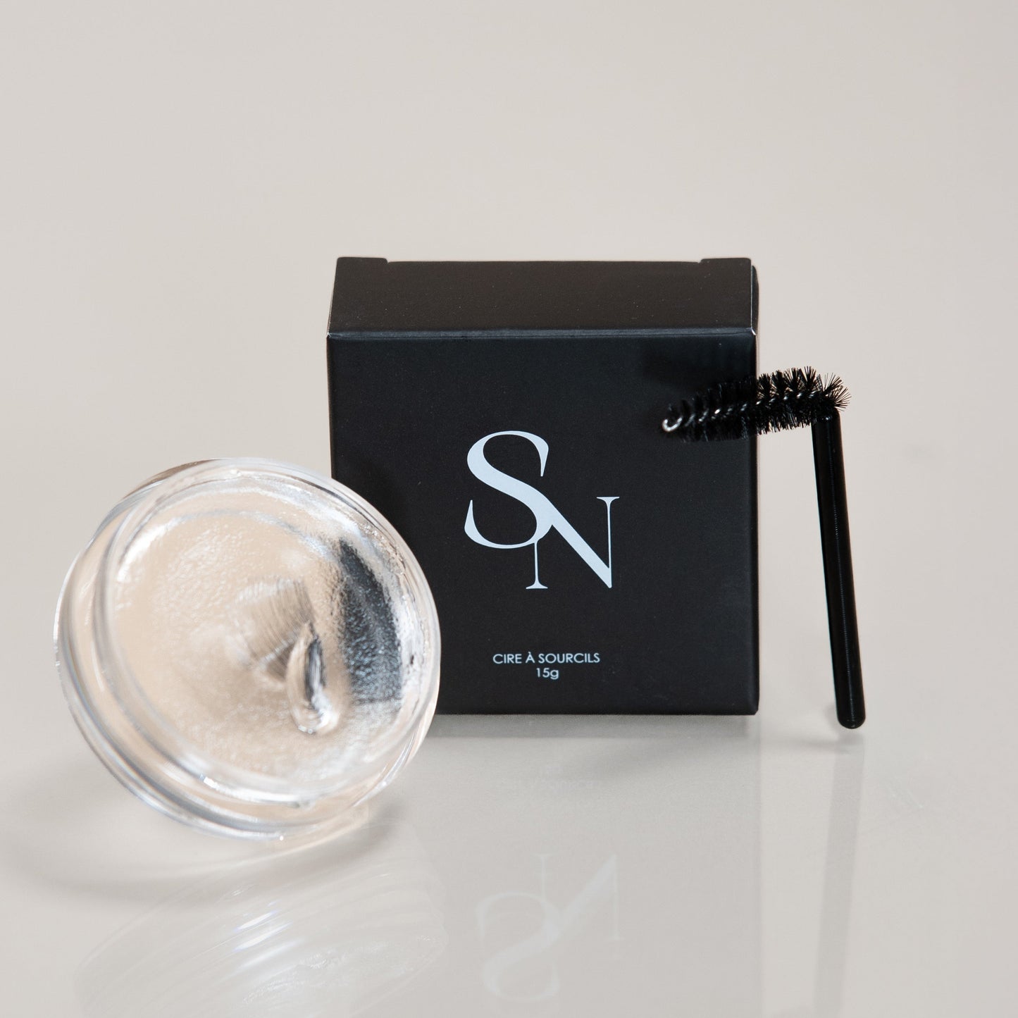 Pot ouvert de cire à sourcils transparente SN BEAUTY, accompagné de sa boîte noire logotée et d’une brosse goupillon noire, sur fond clair.