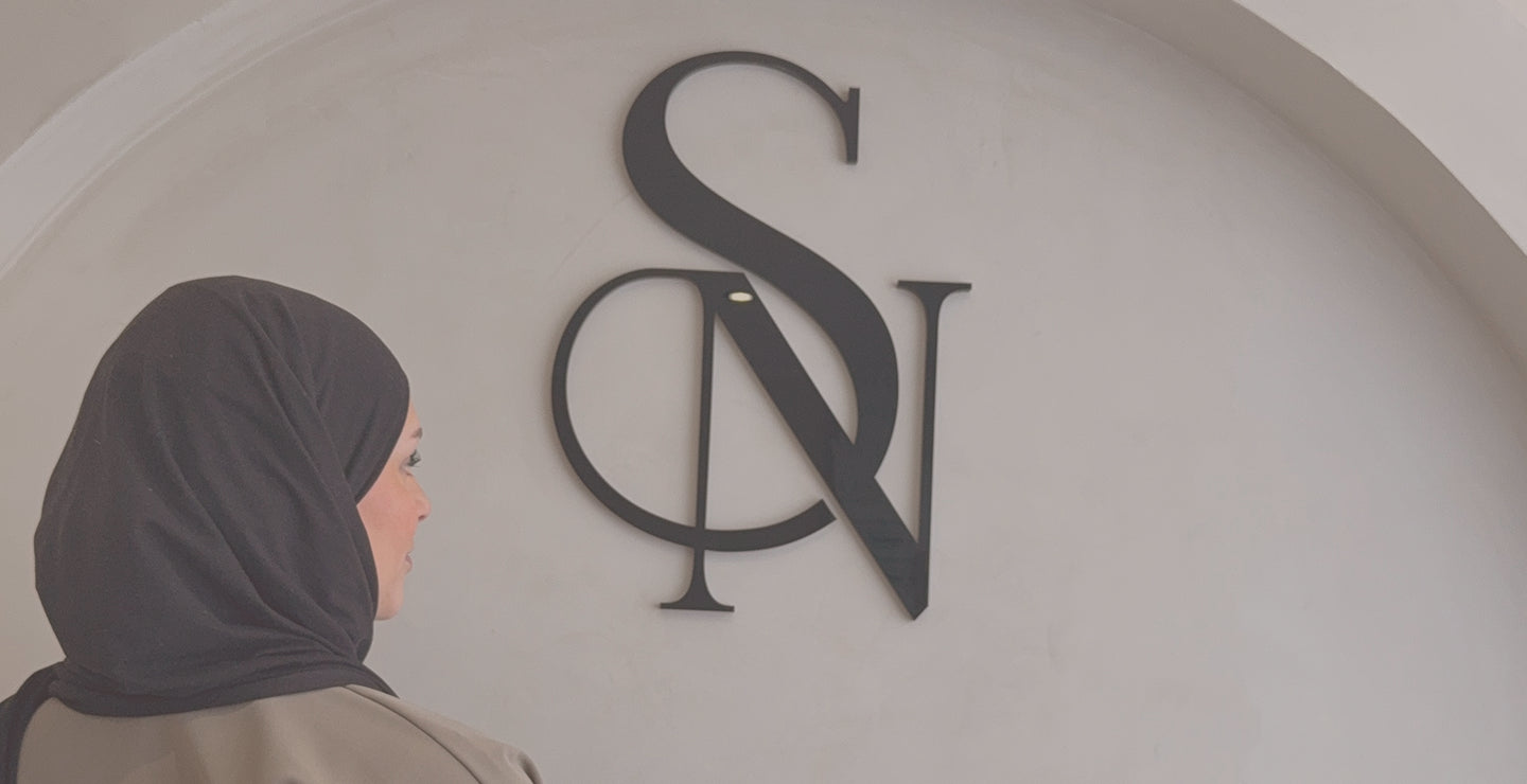 Femme en hijab regardant le logo SN BEAUTY apposé sur un mur clair en arc, ambiance élégante et professionnelle.
