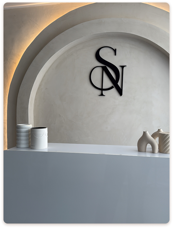Mur en arc avec logo SN Beauté en noir sur fond beige texturé, décor minimaliste et épuré avec vases design.